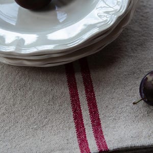 May include: A stack of white ceramic plates with a scalloped edge sits on a folded linen tablecloth with two red stripes.