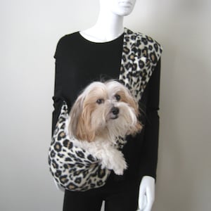 May include: A black and white leopard print dog carrier worn by a person. The carrier is a sling style and is worn over the shoulder. A small white and brown dog is inside the carrier.