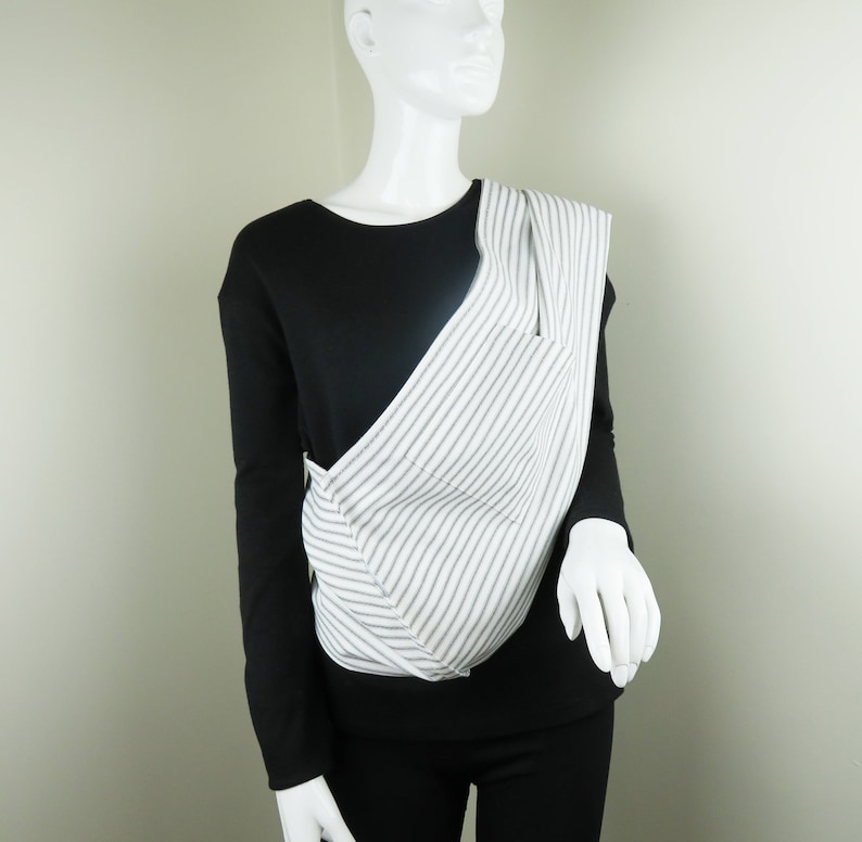 May include: A white and gray striped fabric sling carrier. The carrier is worn diagonally across the body and is made of a soft, comfortable fabric.