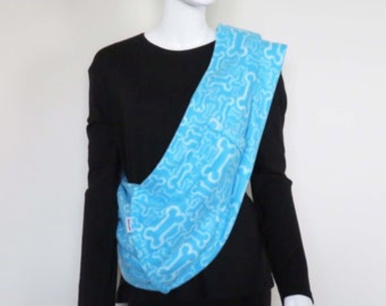 May include: A light blue baby carrier with a white bone pattern. The carrier is being worn by a person wearing a black shirt.