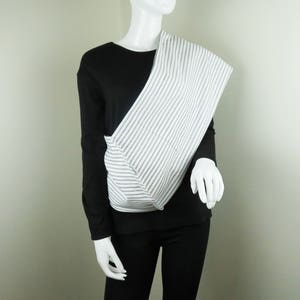 May include: A white and black striped fabric sling worn diagonally across the chest of a mannequin. The sling is made of a soft, woven fabric and has a simple, minimalist design.