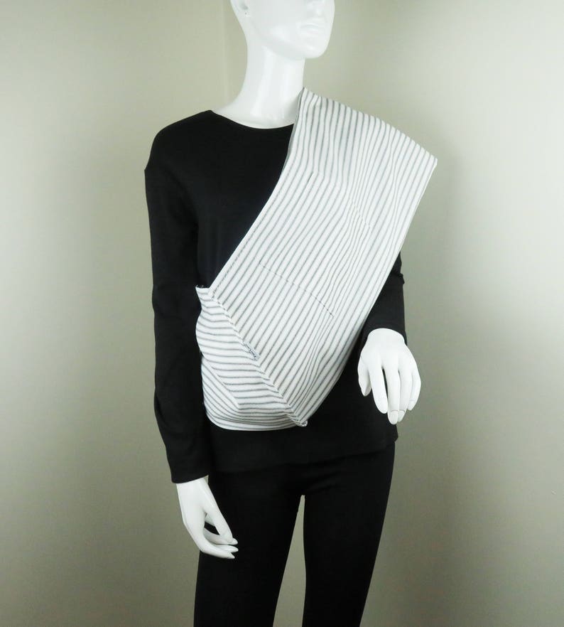 May include: A white and black striped fabric sling worn diagonally across the chest of a mannequin. The sling is made of a soft, woven fabric and has a simple, minimalist design.