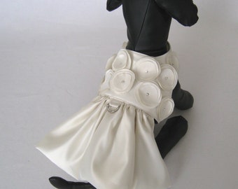 May include: A white satin dog dress with a large flower design and a silver ring detail. The dress is worn by a black dog.