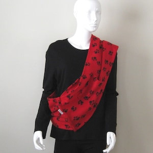 May include: A red fleece pet carrier with black paw prints. The carrier is worn over the shoulder and has a pocket on the front.