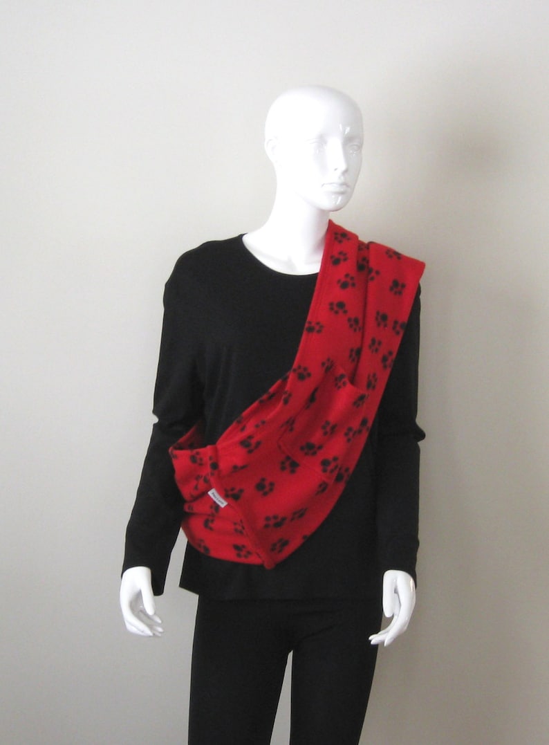 May include: A red fleece pet carrier with black paw prints. The carrier is worn over the shoulder and has a pocket on the front.