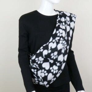 May include: Black and white pet carrier with paw print and heart design. The carrier is worn over the shoulder and has a soft, fleece-like texture.