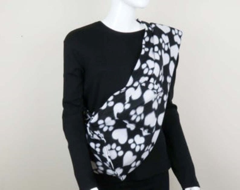 May include: Black and white pet carrier with paw print and heart design. The carrier is worn over the shoulder and has a soft, fleece-like texture.