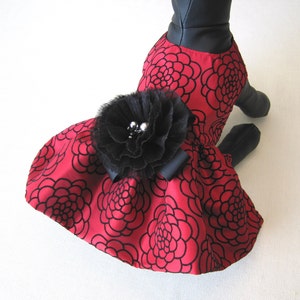 May include: A red dress with a black floral pattern and a black tulle flower with silver embellishments. The dress is designed for a pet.