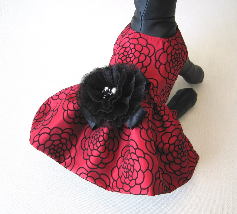 May include: A red dress with a black floral pattern and a black tulle flower with silver embellishments. The dress is designed for a pet.