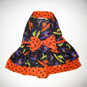 May include: A Halloween-themed dog dress featuring a black background with witch hats in purple, green, and orange. The dress has an orange bow and trim with black star patterns. The collar and hem are trimmed in orange.
