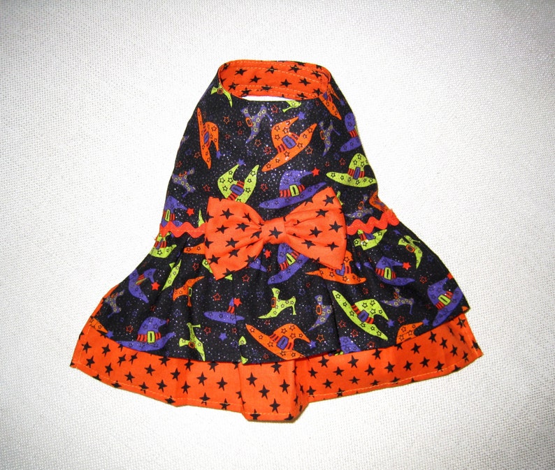May include: A Halloween-themed dog dress featuring a black background with witch hats in purple, green, and orange. The dress has an orange bow and trim with black star patterns. The collar and hem are trimmed in orange.
