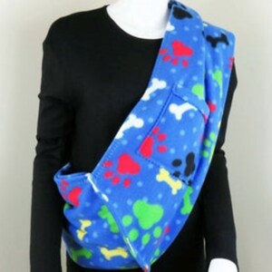 May include: Blue fleece pet carrier with paw print and bone design. The carrier is worn over the shoulder and has a pocket on the front.