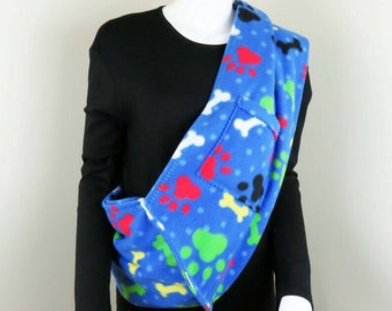 May include: Blue fleece pet carrier with paw print and bone design. The carrier is worn over the shoulder and has a pocket on the front.