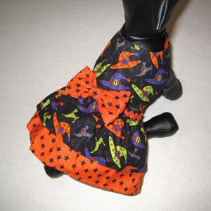 May include: A Halloween-themed dog dress featuring a black fabric with witch hats in orange, purple, and green. The dress has an orange bow and a star-patterned trim. The dress is designed to fit a small dog.