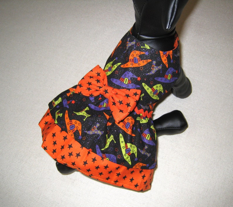 May include: A Halloween-themed dog dress featuring a black fabric with witch hats in orange, purple, and green. The dress has an orange bow and a star-patterned trim. The dress is designed to fit a small dog.