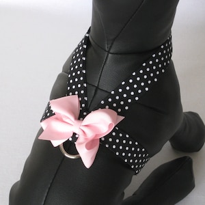 May include: A black and white polka dot dog harness with a light pink bow. The harness has a silver metal ring and is designed to fit around the dog's chest and shoulders. The dog is black.