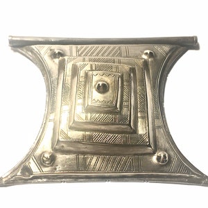 May include: A silver-toned metal decorative piece with a geometric design. The piece has a square shape with a raised center and is decorated with intricate lines and patterns.