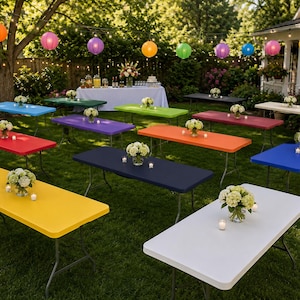 May include: A collection of rectangular folding tables in various solid colors including red, yellow, blue, green, purple, orange, white, and navy, each topped with a floral centerpiece and small candles, arranged across a grassy outdoor lawn.