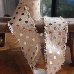 May include: A beige burlap ribbon with gold polka dots and frayed edges. The ribbon is folded and displayed on a wooden surface.