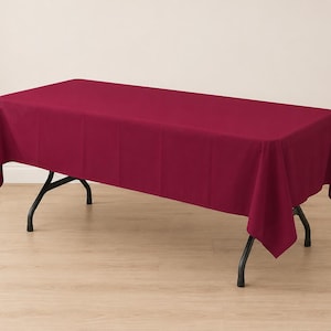 May include: A rectangular, solid deep burgundy tablecloth draped over a folding table with black metal legs. The fabric has a smooth, matte finish and hangs evenly over the edges of the table, covering the top surface and sides completely.