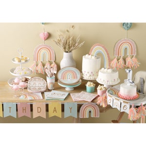 May include: A rainbow-themed first birthday party table setting with a cake, plates, cups, napkins, and a banner that says "Happy Birthday".