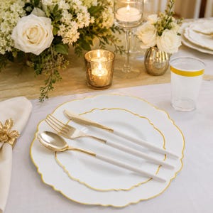 May include: A white plastic dinner plate and salad plate set with scalloped edges and gold trim, accompanied by a matching white plastic fork, knife, and spoon with gold accents, plus a clear plastic cup with a gold rim on a white tablecloth.