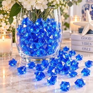 May include: A clear glass vase filled with numerous faceted, royal blue acrylic gems. The vase contains a bouquet of white flowers, and additional blue gems are scattered on the surface around the base of the vase, creating a decorative display.