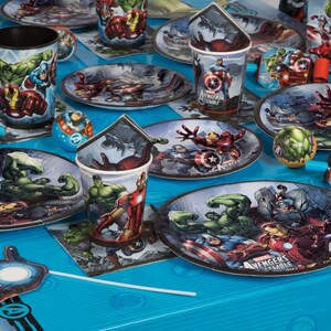 May include: A blue and white themed party table setting with Avengers characters. The table is decorated with plates, cups, napkins, and party favors featuring Iron Man, Hulk, Captain America, and Thor. The text "Avengers Assemble" is printed on some of the plates.