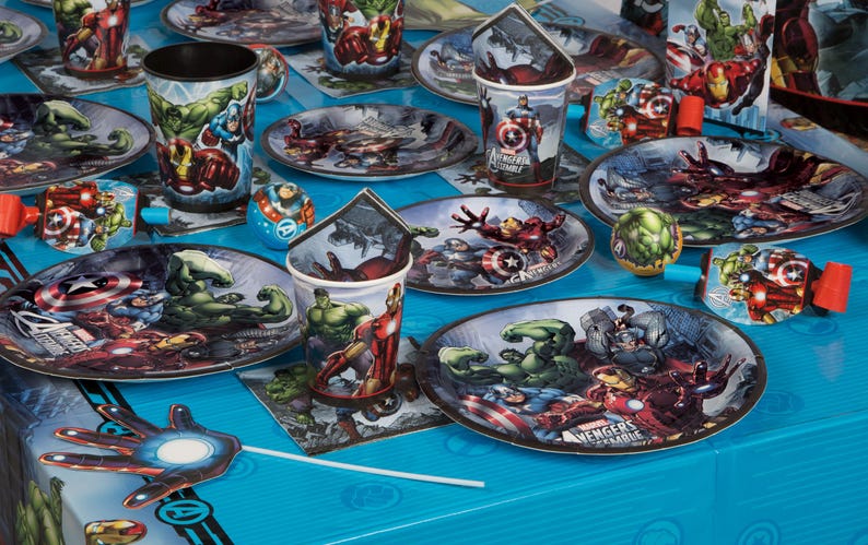 May include: A blue and white themed party table setting with Avengers characters. The table is decorated with plates, cups, napkins, and party favors featuring Iron Man, Hulk, Captain America, and Thor. The text "Avengers Assemble" is printed on some of the plates.