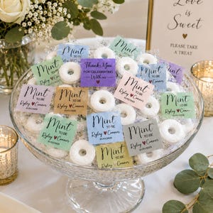 May include: A glass pedestal bowl filled with individually wrapped white ring-shaped mint candies. Each candy features a square paper tag in various pastel colors with the text Mint to be, Ryley heart Conner, and the date 01/01/2025 printed on them.
