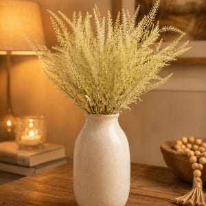 May include: A tall, cylindrical, cream-colored ceramic vase with a smooth, speckled finish. The vase is filled with a dense arrangement of light yellow, feathery, plume-like botanical stems that fan out in a wide, textured, and airy display.