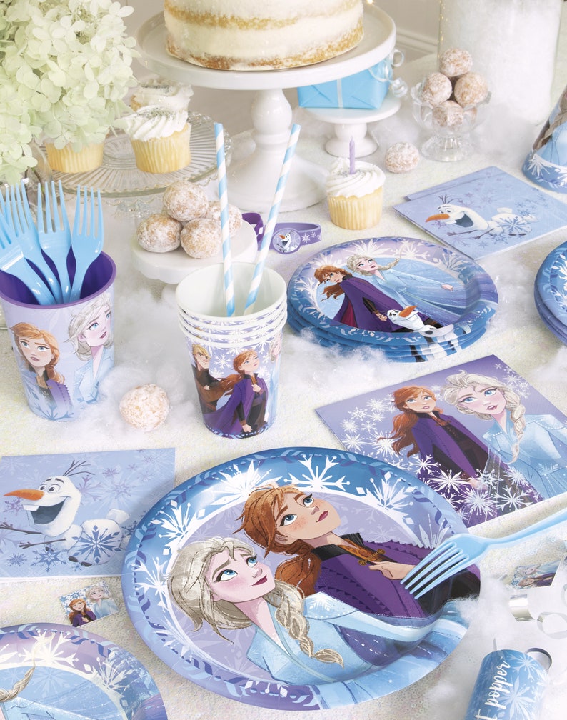 Puede incluir: Una mesa de fiesta con tem&aacute;tica de Frozen. Incluye platos, vasos, servilletas y cubiertos en tonos azules y blancos, adornados con im&aacute;genes de Elsa, Anna y Olaf. La escena est&aacute; decorada con cupcakes, tarta y otras golosinas.