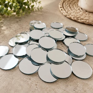 May include: A collection of small, circular, reflective glass mirrors with smooth, polished edges. These individual round mirror pieces are scattered in a pile, each featuring a clear, silver-toned reflective surface that captures light and surrounding details.