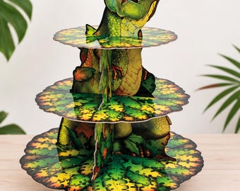 May include: A three-tiered cardboard cupcake stand featuring a green Tyrannosaurus Rex graphic that extends through the center of the circular scalloped shelves. The tiers are decorated with a vibrant jungle leaf pattern in shades of green and yellow.