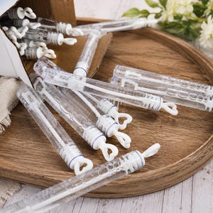 May include: A collection of clear plastic bubble wands with white heart-shaped handles. The wands are cylindrical, filled with bubble solution, and feature a white wand applicator inside. Several wands are scattered, while others remain in a box.