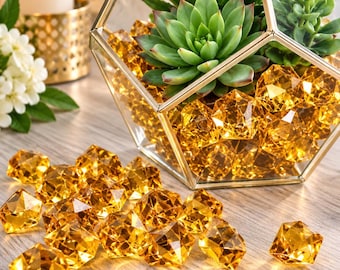 May include: A geometric glass terrarium with a gold metal frame containing green succulent plants and filled with faceted amber-colored acrylic gems. Additional loose amber-colored acrylic gems are scattered in front of the terrarium.