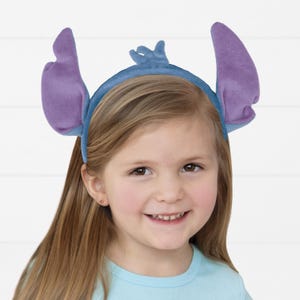Puede incluir: Una diadema azul con orejas de felpa moradas y azules, que se asemejan a un personaje de dibujos animados. La diadema la lleva una ni&ntilde;a con el pelo rubio largo y una camiseta azul claro. La ni&ntilde;a est&aacute; sonriendo.