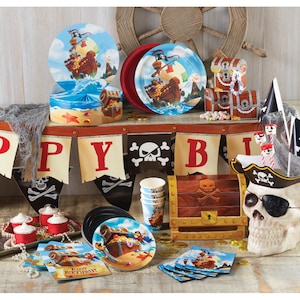 May include: Pirate themed party supplies including plates, cups, napkins, a skull and crossbones banner, a treasure chest, and a pirate skull. The supplies are arranged on a table with a wooden wheel in the background.