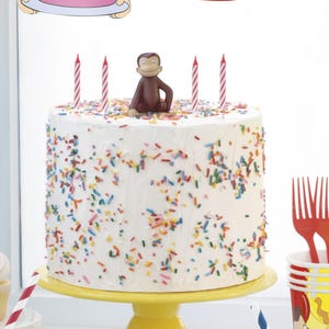 May include: A white frosted cake with colorful sprinkles, topped with a Curious George figurine and red-and-white striped candles. The cake sits on a yellow cake stand, with red forks and party cups nearby. A blue balloon and cupcake are also visible.