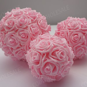 May include: Three pink rose ball ornaments. The balls are made of many small pink roses that are clustered together to form a sphere. The balls are arranged on a white surface.