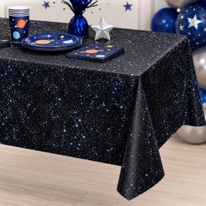 May include: A rectangular plastic tablecloth featuring a dark black space print with scattered white and blue stars. The table is set with matching dark blue paper cups, plates, and napkins, all decorated with colorful planet illustrations.