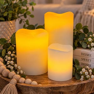 May include: Three cylindrical LED candles with a warm yellow glow and uneven, melted-look tops. The candles are arranged on a round wooden tray alongside a string of light-colored wooden beads and small white flowers with green leaves.