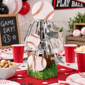 May include: A baseball-themed centerpiece featuring a square base with a green grass print, topped with silver metallic fringe, star cutouts, and circular baseball-patterned cardstock accents. The design includes a large baseball sphere on top.