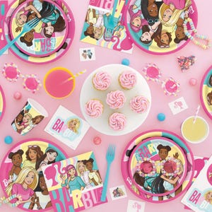 May include: Pink and yellow Barbie themed party supplies, including plates, napkins, cups, and cupcake wrappers. The plates feature a group of diverse Barbie dolls with the text "Barbie" and "Girls" on them. The napkins have a pink and yellow design with the text "Barbie" on them. The cups are pink and have a white rim. The cupcake wrappers are pink and have a white rim. The cupcakes are pink and have white frosting.