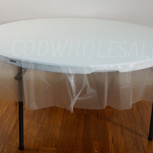 May include: A clear plastic table cover with a white tablecloth underneath. The table cover is draped over a round table with black legs. The table cover is designed to protect the tablecloth from spills and stains.