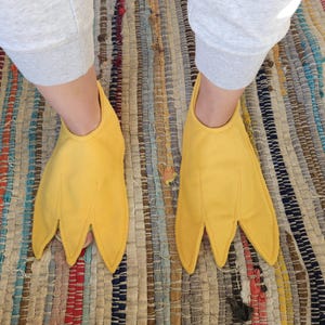 Puede incluir: Un par de pantuflas de tela de color amarillo brillante con forma de patas de ave palmeadas. Las pantuflas presentan tres dedos puntiagudos distintos con detalles de costura, cubriendo el empeine y extendiéndose más allá de los dedos.