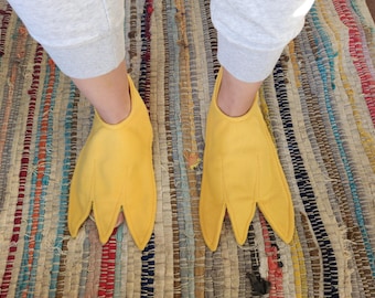 Puede incluir: Un par de pantuflas de tela de color amarillo brillante con forma de patas de pato palmeadas. Las pantuflas presentan tres dedos puntiagudos distintos con detalles de costura, cubriendo la parte delantera de los pies y dejando el talón abierto.