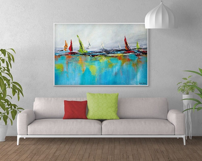 May include: Abstract painting of sailboats on a blue and green water surface. The painting is framed in a white frame and is hanging on a white wall above a white couch with red and green pillows.