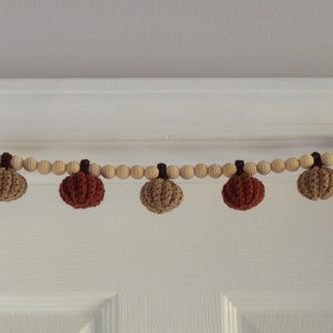 May include: A fall-themed garland featuring small, crocheted pumpkins in shades of brown and orange. The pumpkins are strung along a wooden bead strand, creating a rustic and seasonal decorative piece. The garland is perfect for autumn decor.