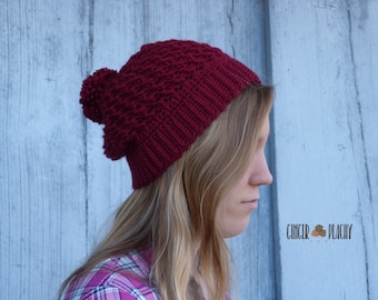 May include: A burgundy knit hat with a textured pattern and a pom-pom. The hat is made of a soft, warm material, perfect for cold weather. The hat is shown on a person. The shop name "Ginger Peachy" is in the bottom right corner.